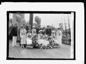 Navy Sailors and women in dress and large hats pose with flowers on a ship.